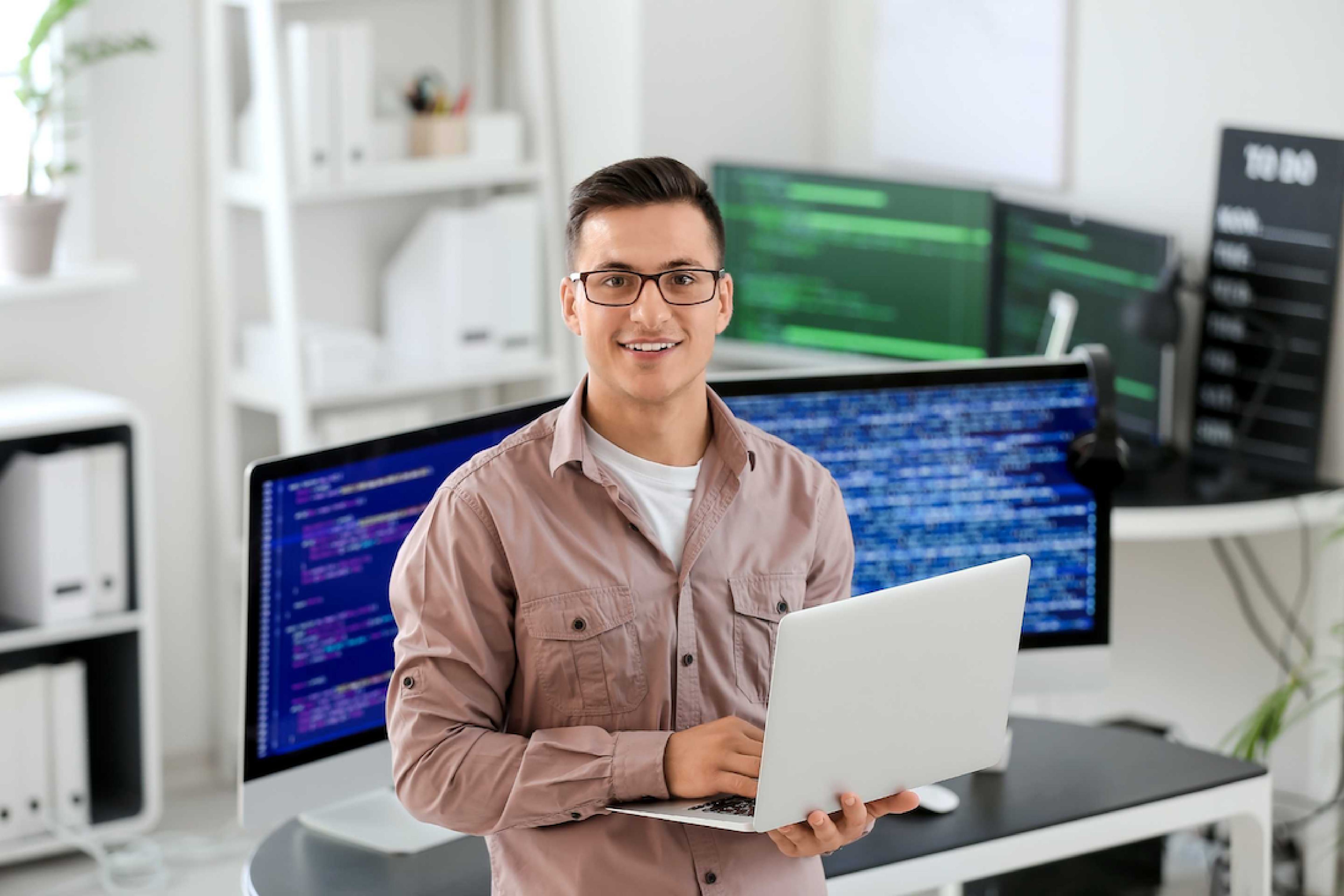 Jonge man met bril houdt laptop in handen en op de achtergrond meerdere computerschermen