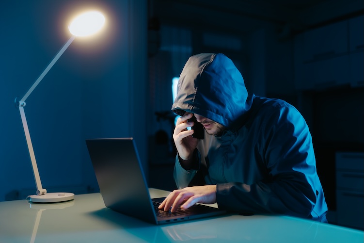 In a dark room, a hacker works on his laptop, talking on the phone and celebrating success after achieving fraudulent goals in the digital world.